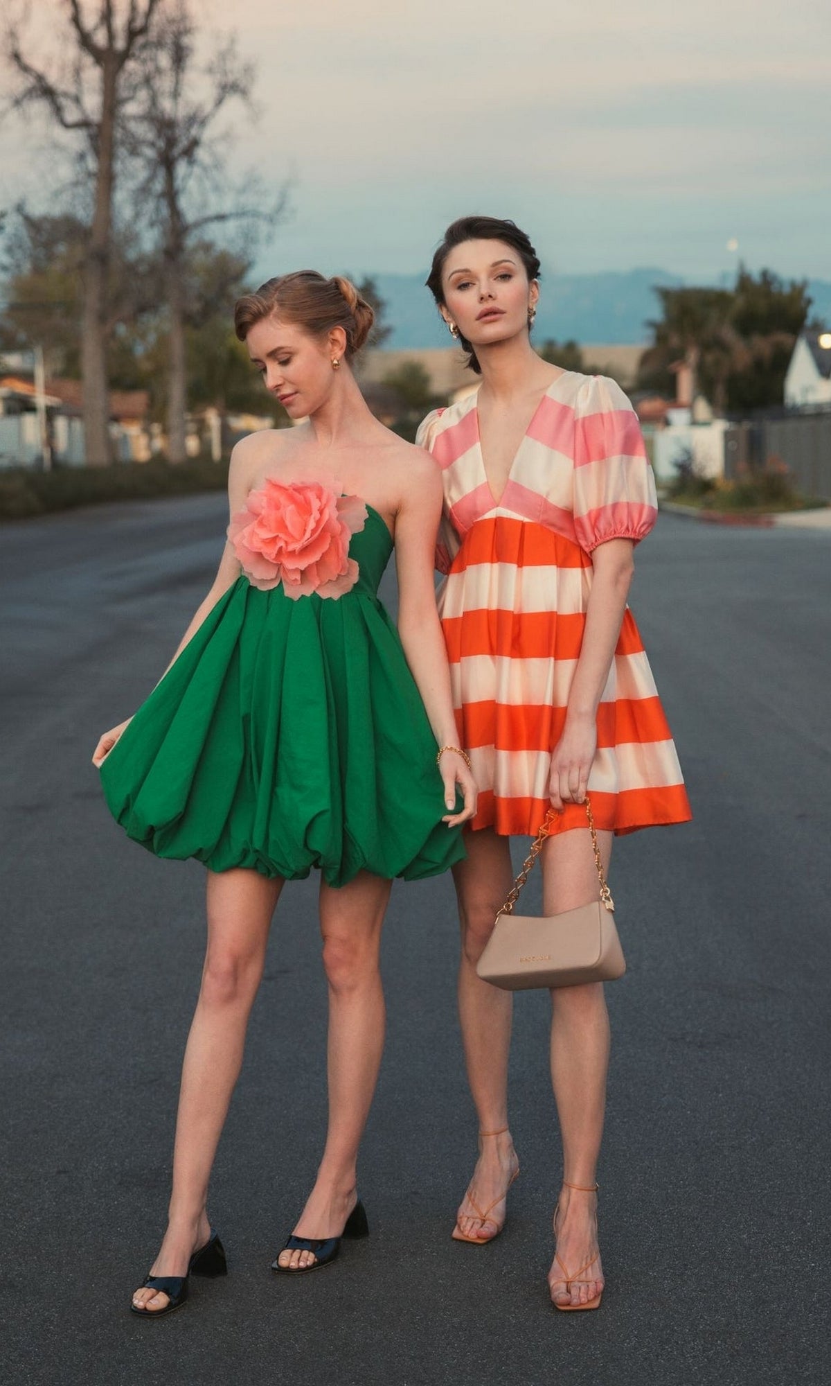 Two women stand on a street; one wears the Mac Duggal 20931 Short Bubble Party Dress with a large pink flower, while the other sports a pink and orange striped semi-formal dress with puff sleeves, carrying a beige handbag.